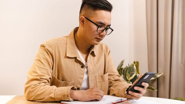 Homem consultando o celular e fazendo anotações à mesa
